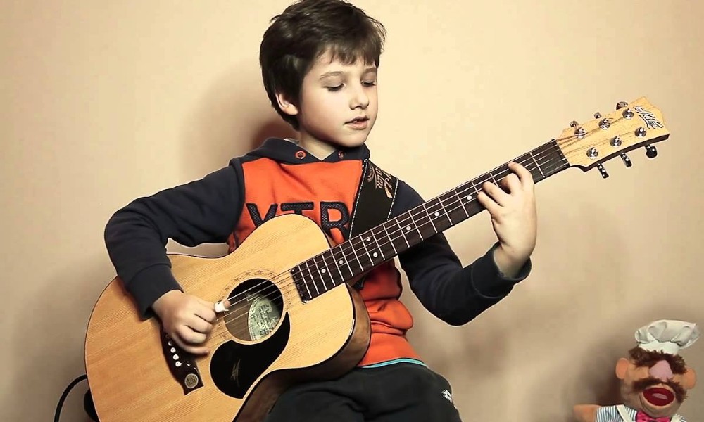 An image representing a small boy sitting and playing Guitar and a Doll next to him